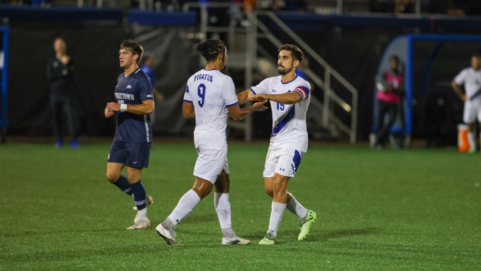 Two Seton Hall men's soccer teammates stand on the field during a play in a game against the No. 1 Georgetown Hoyas.